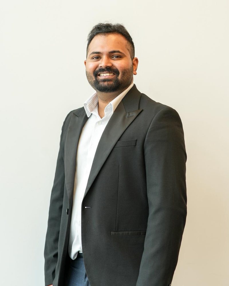 Thomas Kunjumon, smiling, in a dark jacket and white shirt against a plain wall